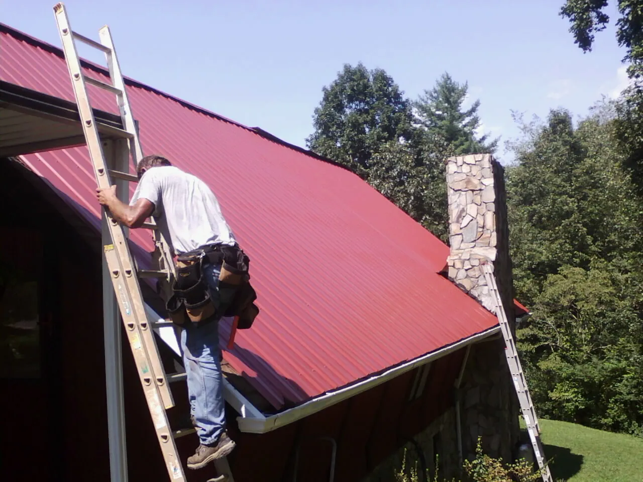 Roofer on a ladder beside a red metal roof and stone chimney — Soffit & Fascia Repair by Roofing Brothers in Canton Center